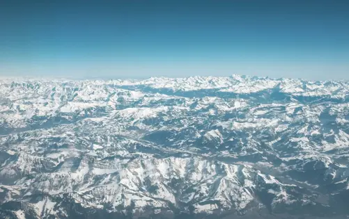 Vue aérienne d’une vaste chaîne de montagnes enneigées, avec d’innombrables sommets s’étendant à perte de vue sous un ciel bleu clair.
