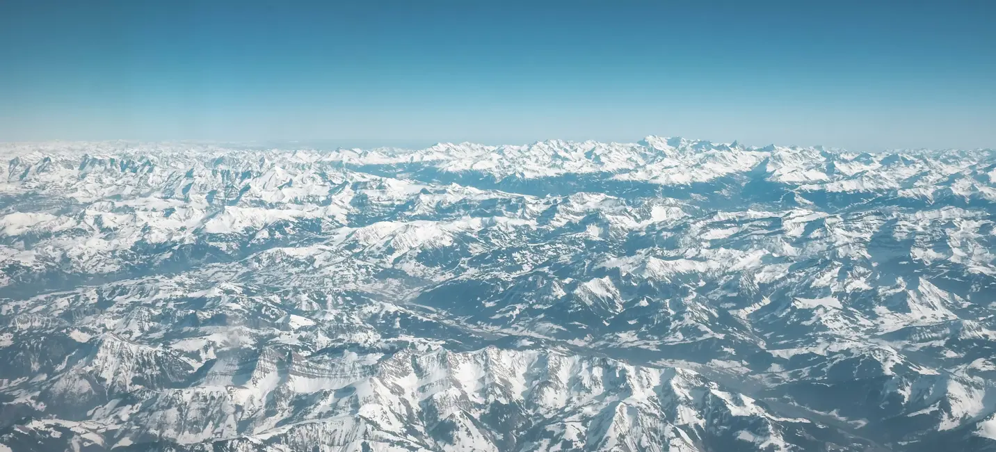 Вид с воздуха на обширный заснеженный горный хребет, бесконечные вершины которого тянутся вдаль под ясным голубым небом.