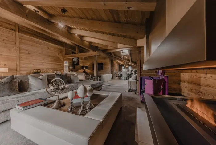 Luxurious chalet-style living room with exposed wooden beams, plush velvet sofas, elegant decor, and a modern fireplace creating a warm and cozy mountain ambiance.