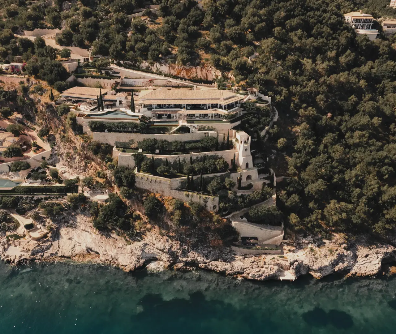 Aerial view of a luxury coastal villa built into a hillside with terraces, gardens, and a pool overlooking the turquoise sea.