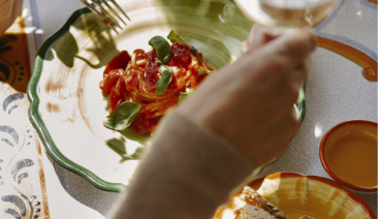Person enjoying a plate of pasta with tomato sauce and basil, served with bread and a glass of white wine.