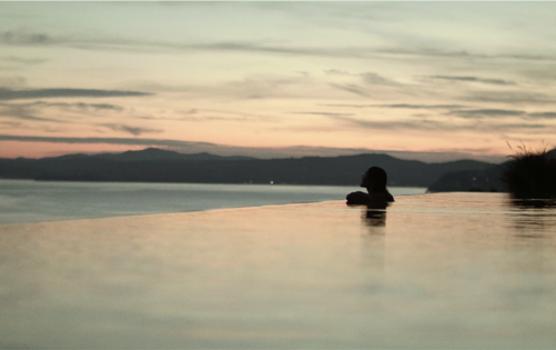 Silhouette d’une personne se détendant dans une piscine à débordement au coucher du soleil, face à la mer calme et aux montagnes lointaines.