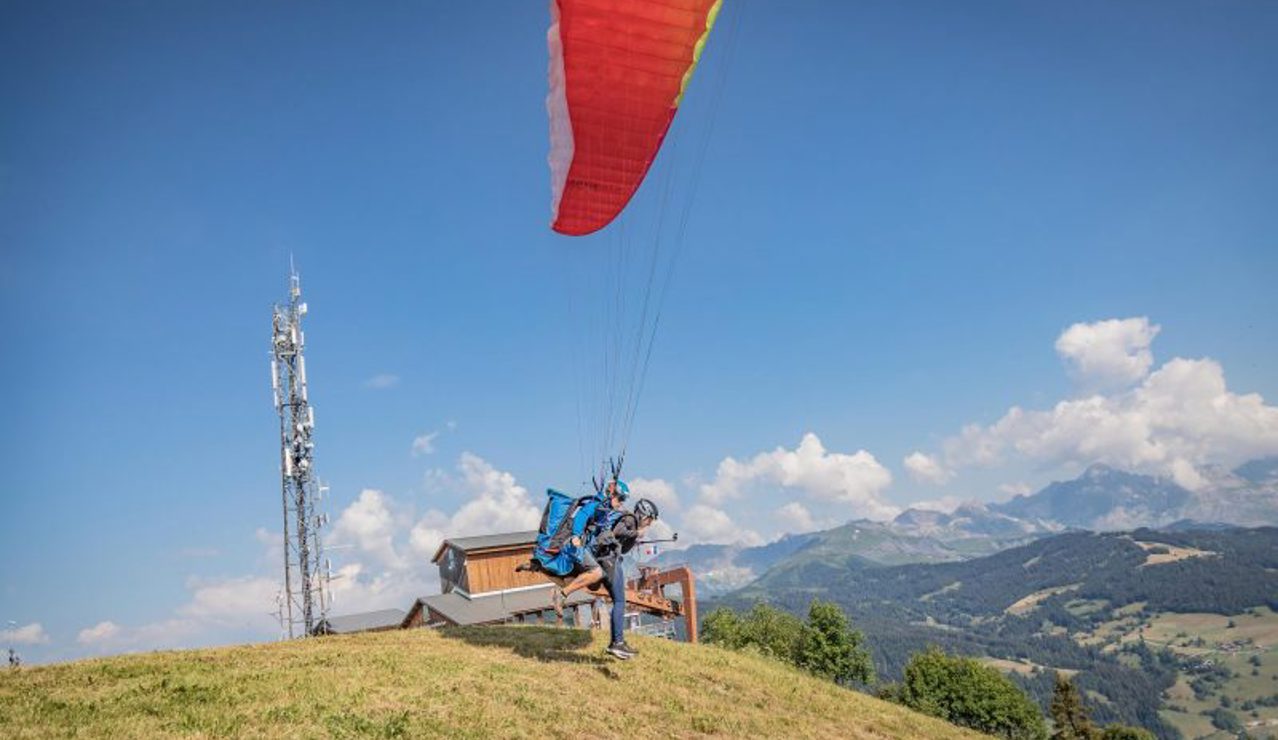 Paraglider taking off from a mountain slope with a red canopy and scenic alpine landscape in the background.