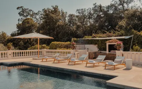 Terrasse élégante au bord de la piscine de style méditerranéen avec transats, parasols blancs et cuisine extérieure entourée de verdure sous un ciel clair.
