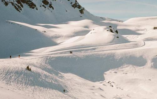Skiers gliding across snowy alpine slopes beneath rugged mountain peaks in bright daylight.