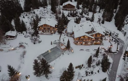 Vue aérienne d’un domaine alpin exclusif entouré de forêts de pins enneigées, mettant en valeur une architecture en bois élégante, un éclairage extérieur chaleureux et une piscine extérieure chauffée dans un resort privé de montagne.
