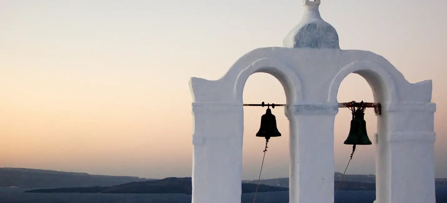 Clocher blanchi à la chaux surplombant la mer Égée au coucher du soleil, sous un ciel pastel avec des îles lointaines à l’horizon.