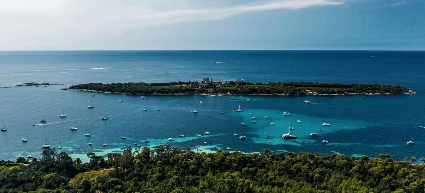 Aerial view of a Mediterranean island surrounded by turquoise waters and anchored yachts.