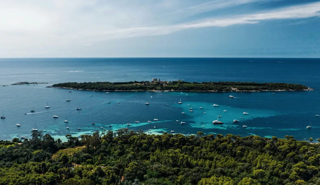 Aerial view of a Mediterranean island surrounded by turquoise waters and anchored yachts.
