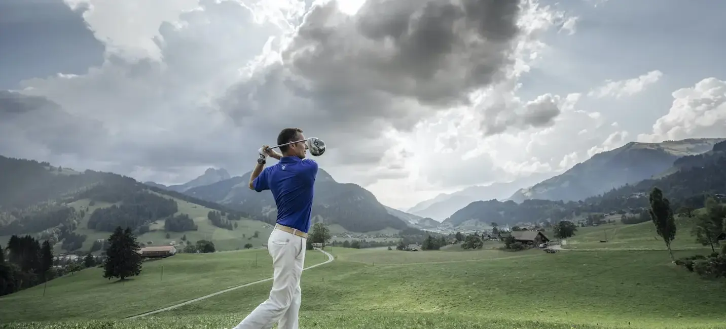 Гольфист делает удар на зеленом альпийском поле, окружённом горами под выразительным небом.