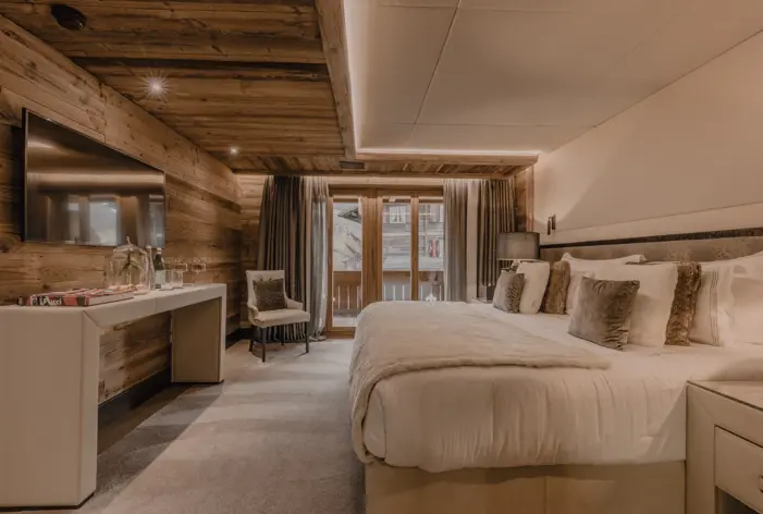 A cozy and elegant chalet bedroom with a large bed dressed in soft white linens and fur accents. The space features warm wood-paneled walls, ambient lighting, and a private balcony visible through glass doors, creating a serene mountain retreat.