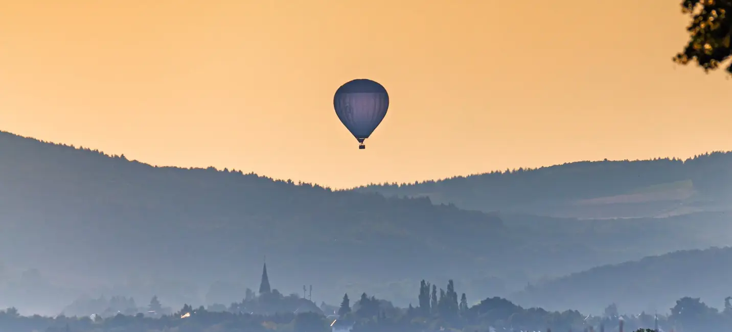 A hot air balloon drifts above a valley at sunrise, with soft golden light over distant hills and villages.