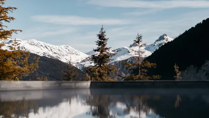 Snow-covered alpine mountains reflected in a still outdoor pool surrounded by pine trees.