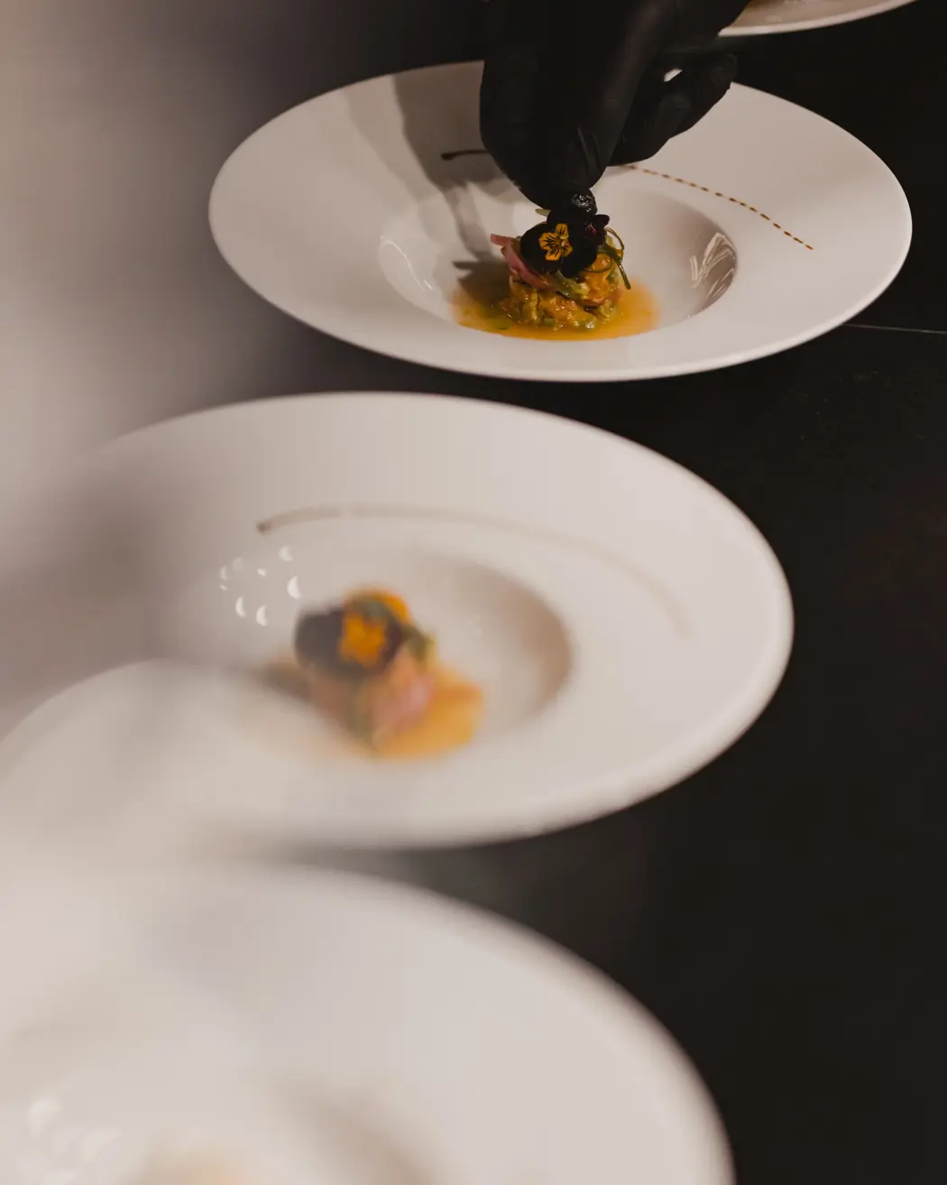 Main du chef dressant un plat gastronomique avec précision, garni de fleurs comestibles sur une assiette blanche.