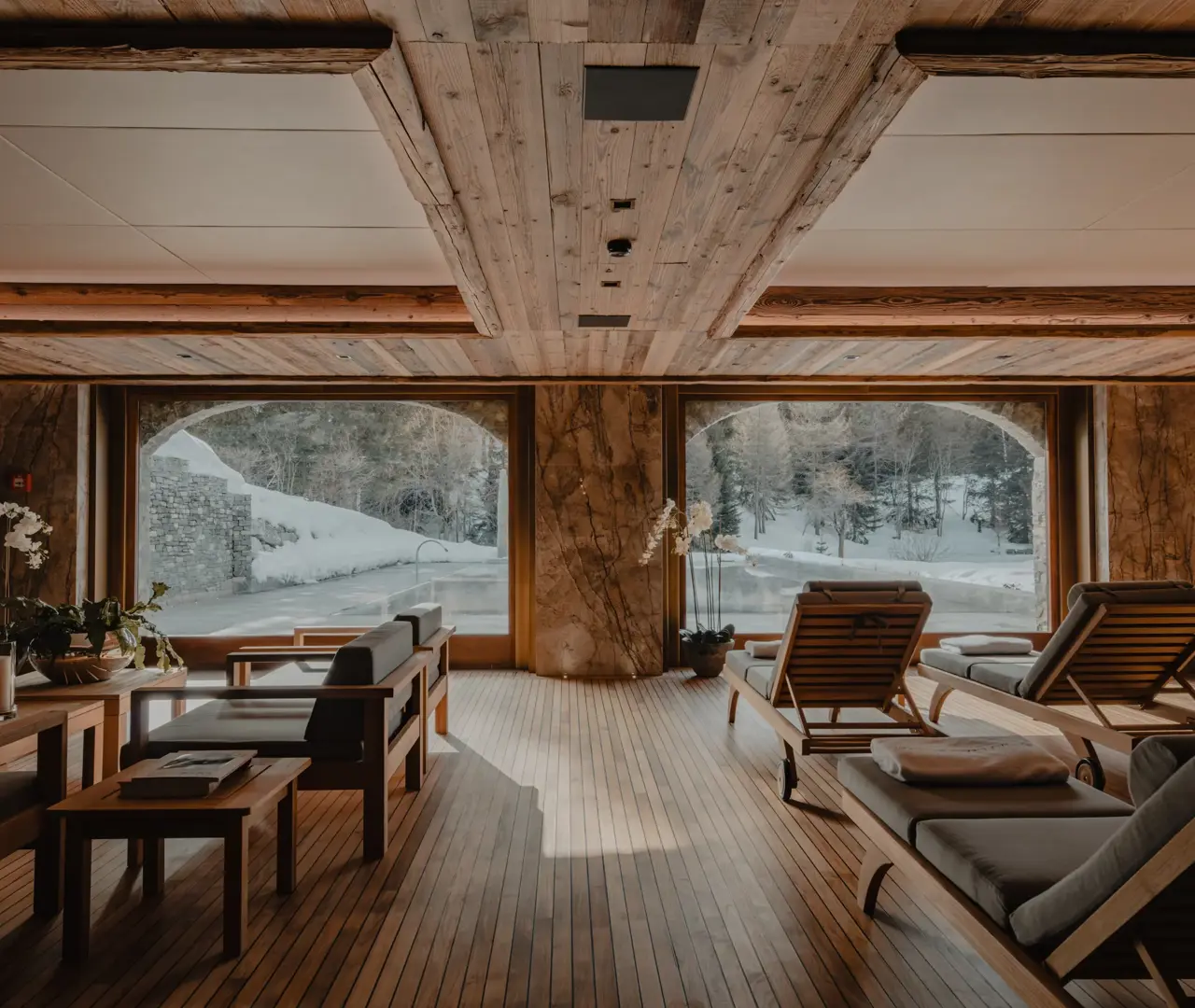 Elegant alpine spa lounge with wooden interiors, large windows overlooking snowy mountains, and cozy relaxation chairs.
