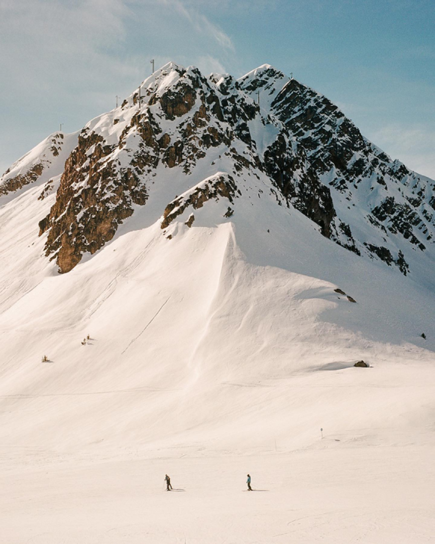 Two skiers on snowy slopes beneath a rugged mountain peak under a clear blue sky.