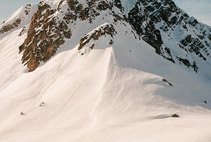 Two skiers on snowy slopes beneath a rugged mountain peak under a clear blue sky.