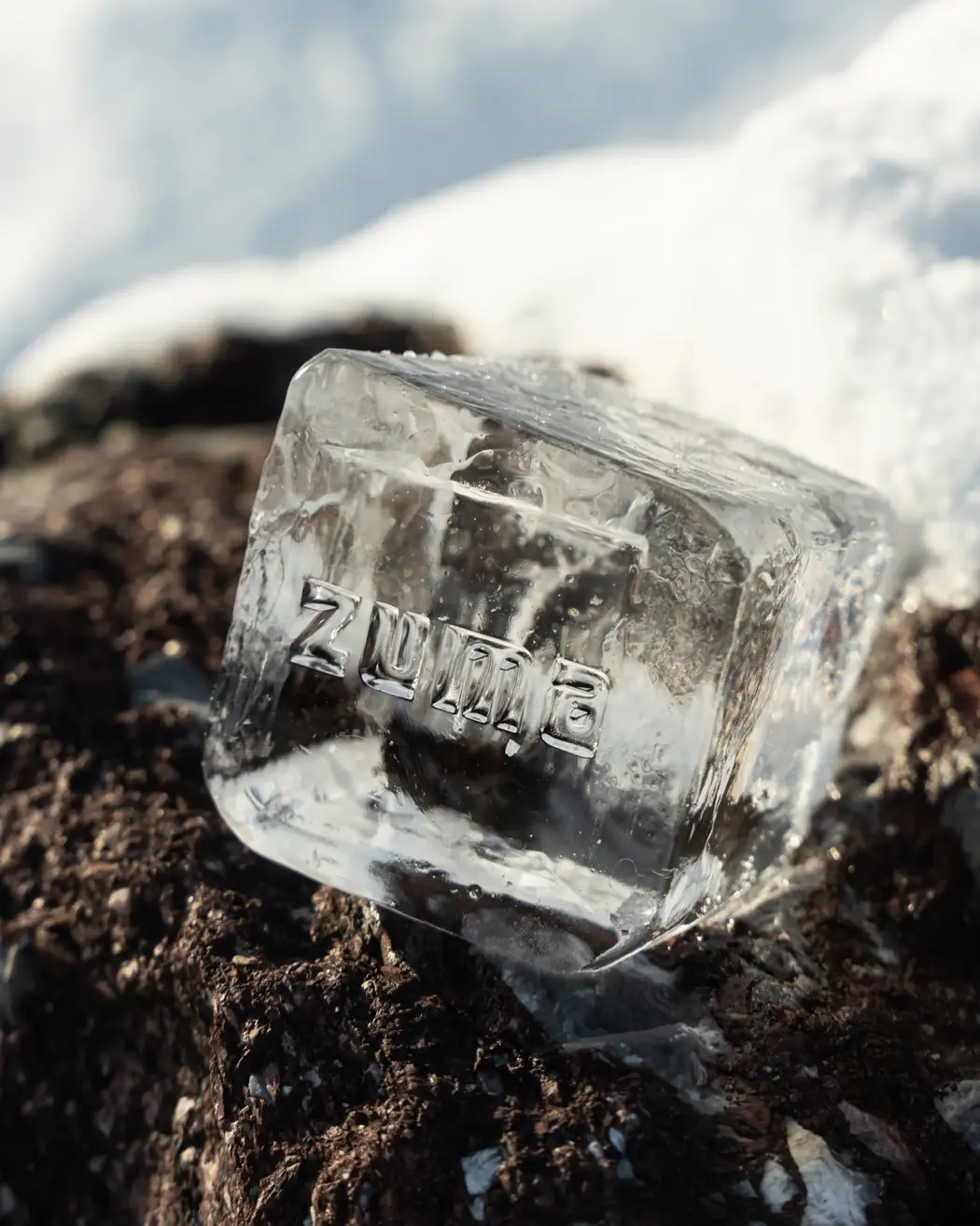 Clear ice cube with the word “Zuma” engraved, resting on snow-covered ground under sunlight.