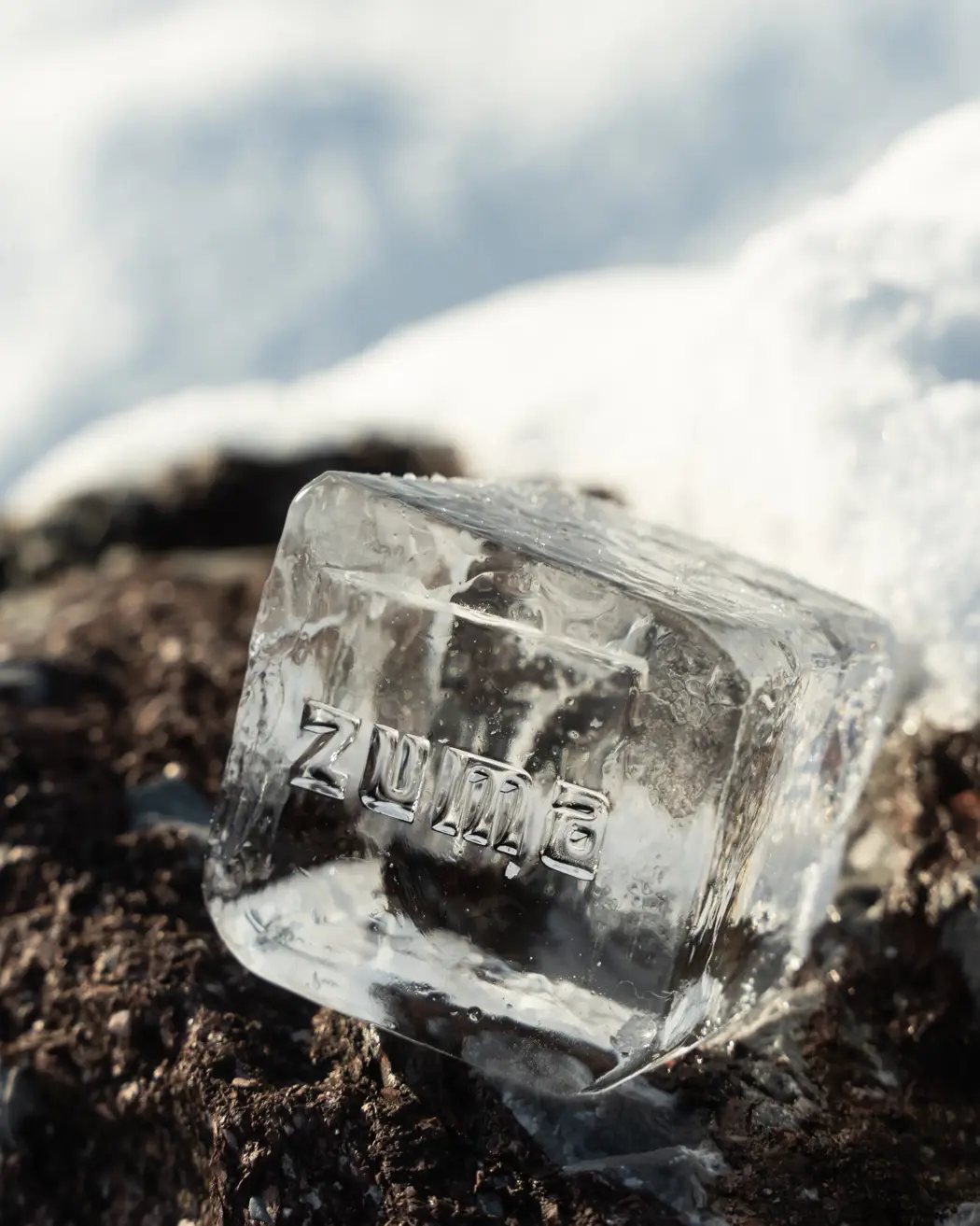 Clear ice cube with the word “Zuma” engraved, resting on snow-covered ground under sunlight.