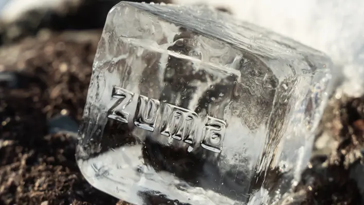 Clear ice cube with the word “Zuma” engraved, resting on snow-covered ground under sunlight.