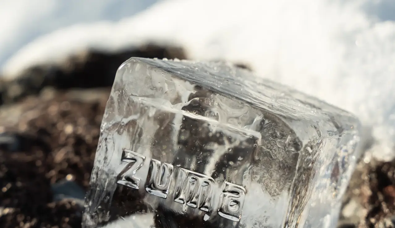Clear ice cube with the word “Zuma” engraved, resting on snow-covered ground under sunlight.