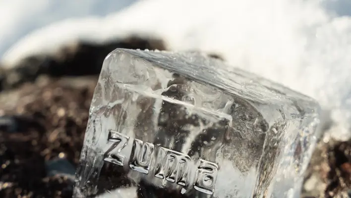 Clear ice cube with the word “Zuma” engraved, resting on snow-covered ground under sunlight.
