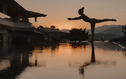 Silhouette d’une femme pratiquant le yoga dans une posture d’équilibre au bord d’une piscine à débordement au coucher du soleil, avec les montagnes en arrière-plan.