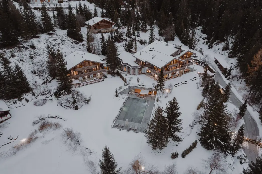 Aerial daytime view of a luxury alpine chalet estate nestled in a snowy mountain landscape, featuring two interconnected lodges, a heated outdoor pool, and surrounding pine forest in an exclusive ski resort.