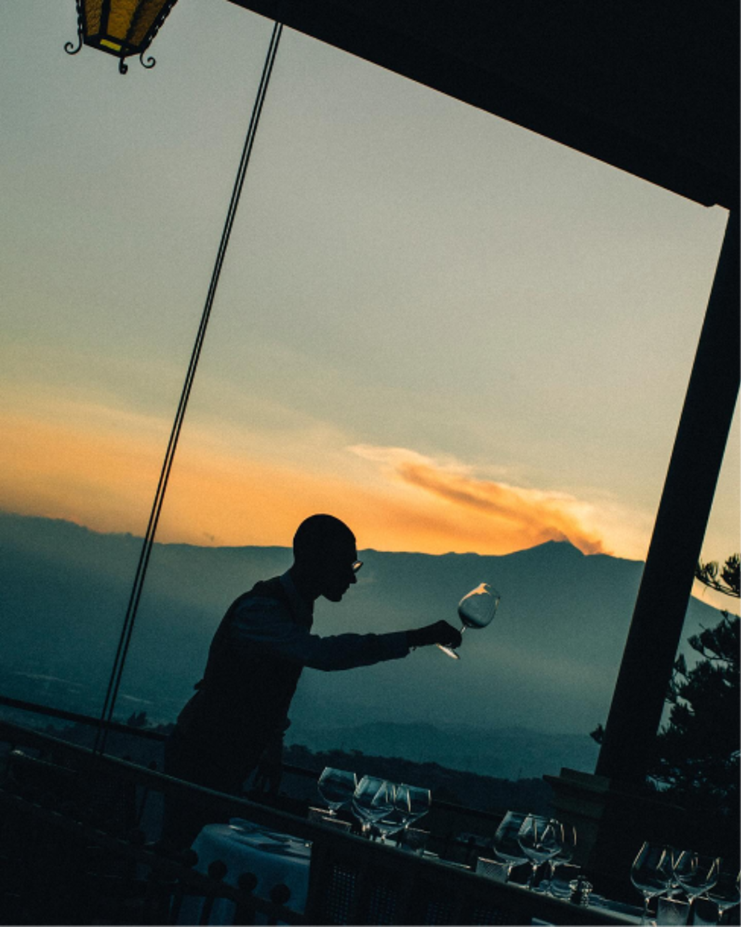 Silhouette of a person pouring wine at sunset on a terrace overlooking distant mountains.