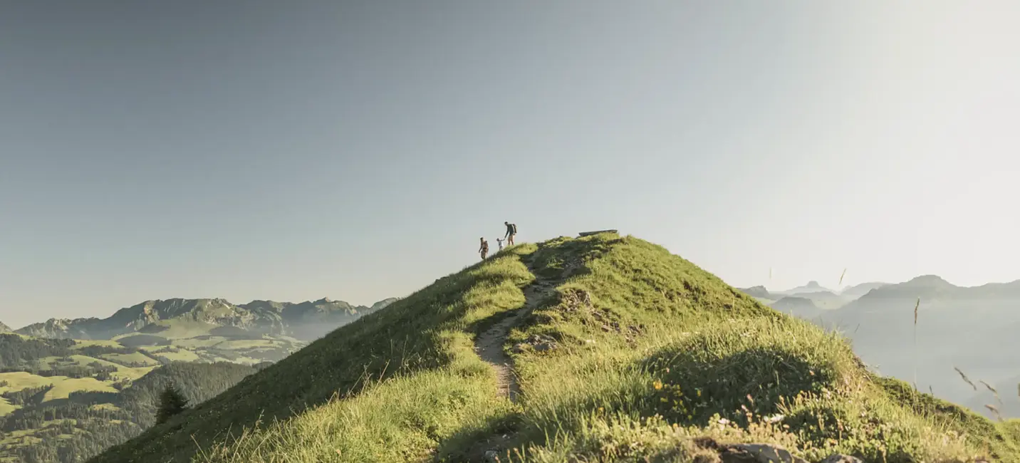 Hikers walk along a grassy alpine ridge under a clear sky, surrounded by panoramic mountain views.