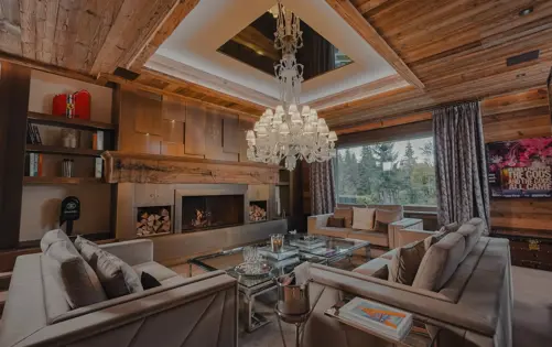 Salon élégant d’un chalet alpin de luxe, avec un grand lustre en cristal, une cheminée moderne, des canapés en velours et une vue panoramique sur la forêt — une alliance parfaite entre chaleur et sophistication alpine.