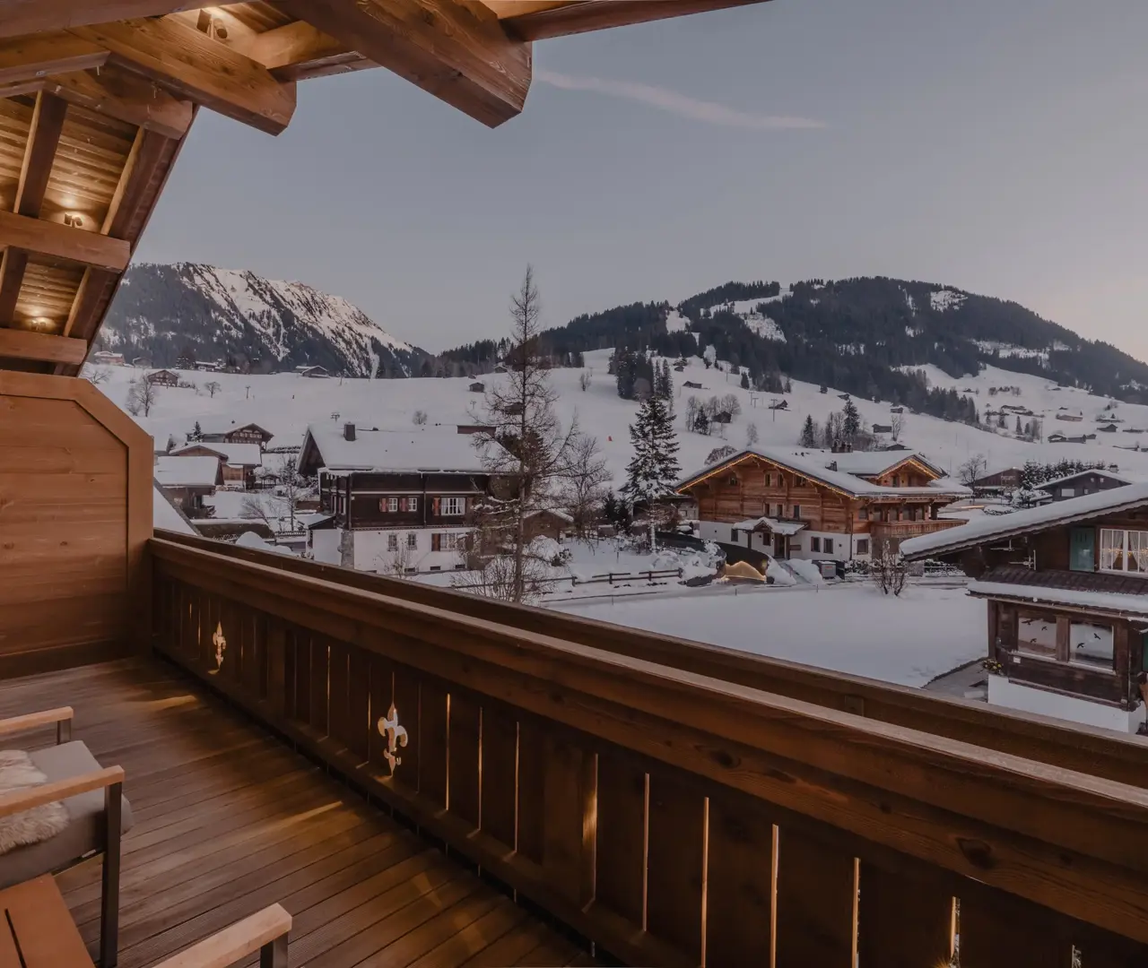 Chalet balcony view at dusk overlooking snowy alpine rooftops and mountain slopes.
