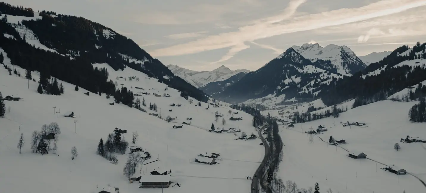 Vallée alpine enneigée avec des chalets en bois dispersés, une route sinueuse et des sommets montagneux sous un ciel d’hiver doux.