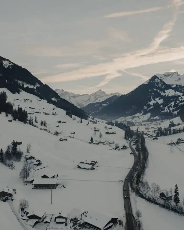 Vallée alpine enneigée avec des chalets en bois dispersés, une route sinueuse et des sommets montagneux sous un ciel d’hiver doux.