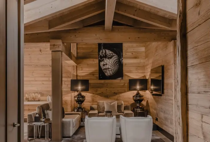 Salon chaleureux et élégant de style chalet avec murs en bois clair et poutres apparentes, mobilier moelleux, lampes sombres et un portrait en noir et blanc en point focal.
