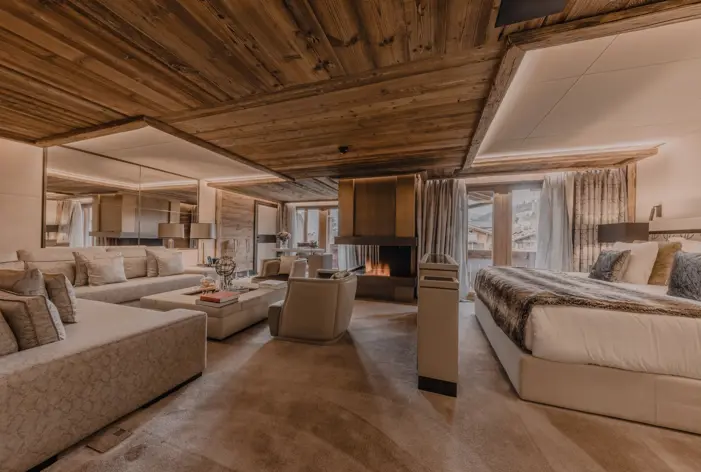 A luxurious open-plan suite with a seamless blend of bedroom and living spaces. The interior features warm wood paneling, plush beige seating, and a modern central fireplace that adds both warmth and ambiance. Natural light pours through the large windows, highlighting the elegant mix of textures — from the fur throw on the bed to the mirrored wall and soft carpeting — creating a refined alpine retreat.