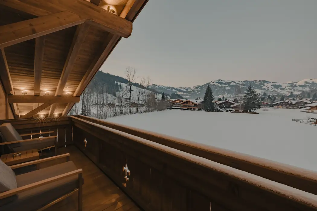 Balcon en bois chaleureusement éclairé avec des chaises longues confortables donnant sur un village alpin enneigé au crépuscule, entouré de montagnes et de pins.
