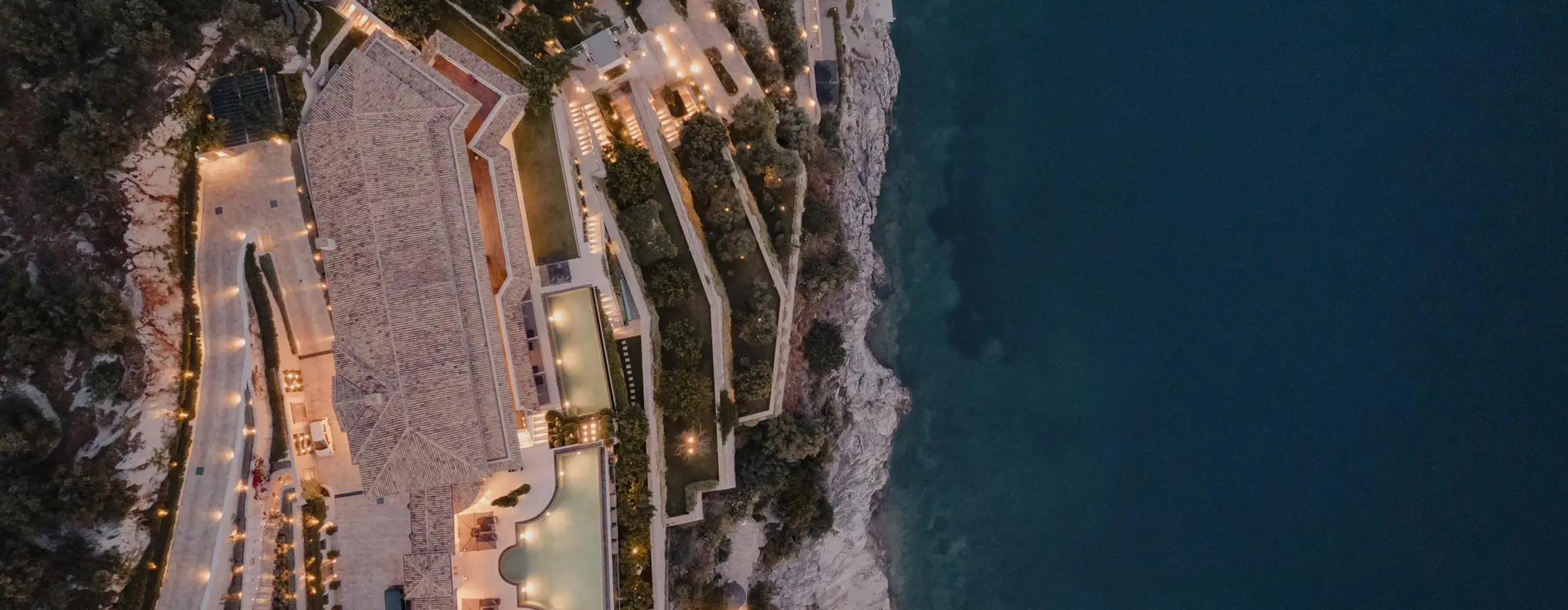 Aerial view of a luxurious cliffside resort at dusk, with illuminated terraces, pools, and pathways overlooking a calm blue sea.