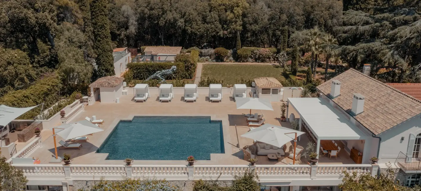 Une villa luxueuse avec une grande piscine rectangulaire entourée de transats et de parasols, située sur une terrasse donnant sur une végétation dense. La propriété présente une architecture de style méditerranéen avec des murs blancs, des toits en tuiles et un coin salon extérieur ombragé.
