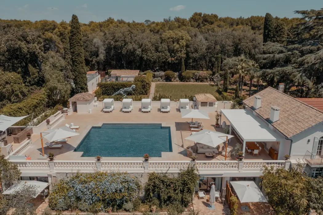 فيلا فاخرة تحتوي على مسبح مستطيل كبير محاط بكراسي استلقاء ومظلات، تقع على شرفة تطل على أشجار كثيفة. يتميز العقار بطراز معماري متوسطي بجدران بيضاء وأسقف من القرميد ومكان جلوس خارجي مظلل.