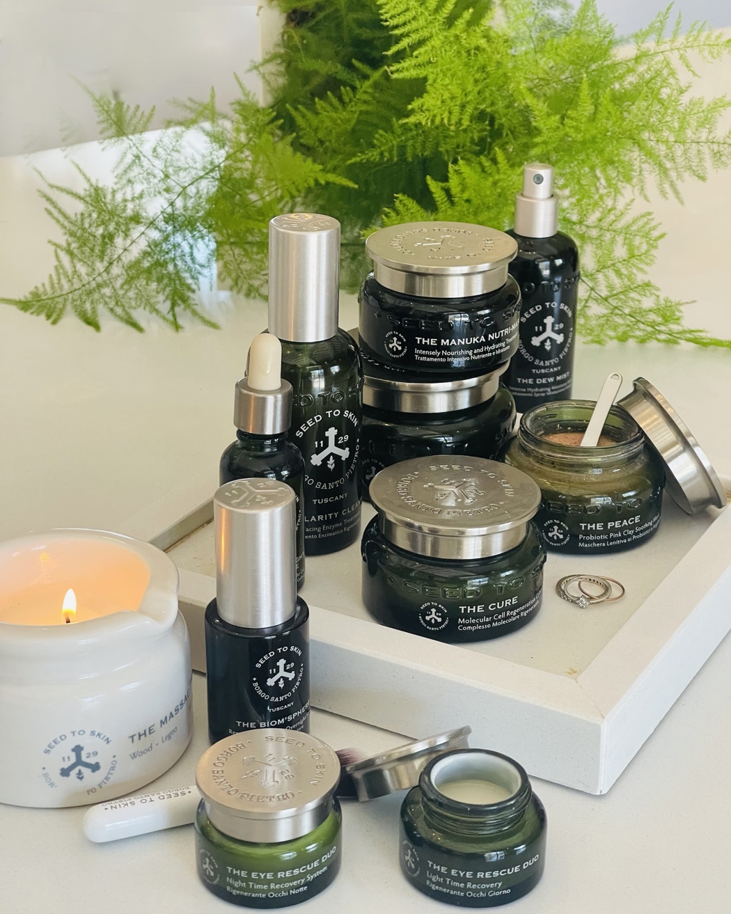 Collection de produits de soin Seed to Skin comprenant des pots, des pipettes et une bougie allumée disposés sur un plateau blanc avec des plantes vertes en arrière-plan.