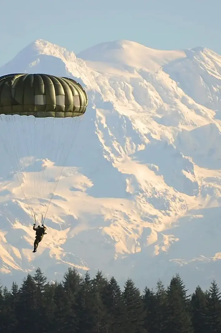 A lone parachutist descends through clear alpine air with snow-capped peaks rising in the distance — an image of freedom, precision, and mountain adventure.