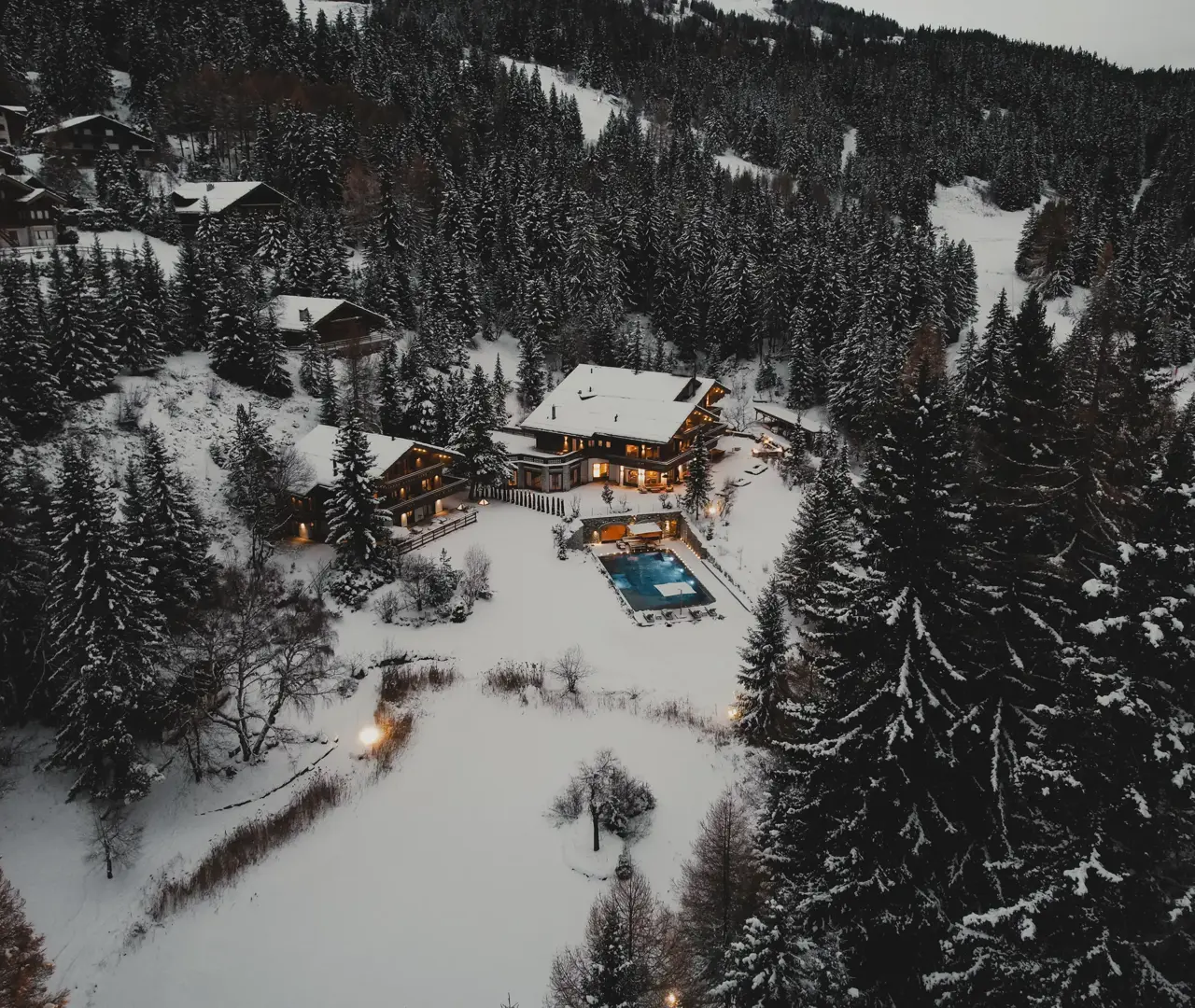 Vue aérienne d’un chalet de luxe entouré de forêts de pins enneigées, avec piscine extérieure chauffée et terrasses illuminées, situé dans un domaine alpin exclusif au crépuscule.