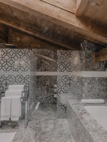 A stunning alpine bathroom featuring a sloped wooden ceiling, elegant grey marble surfaces, and decorative glass panels with intricate geometric patterns. The bathtub and vanity blend modern luxury with rustic charm, enhanced by warm lighting and natural textures.