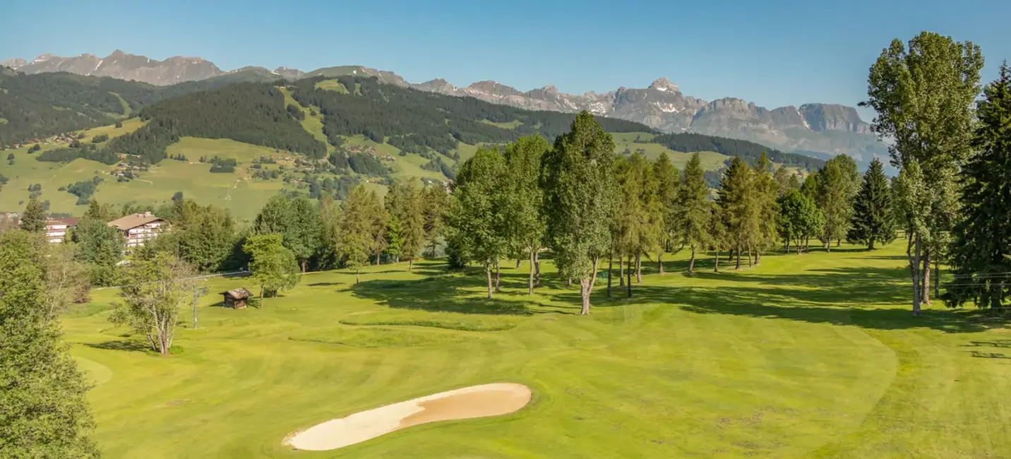 Scenic golf course surrounded by lush greenery and trees with mountains in the background under a bright blue sky in Megève.
