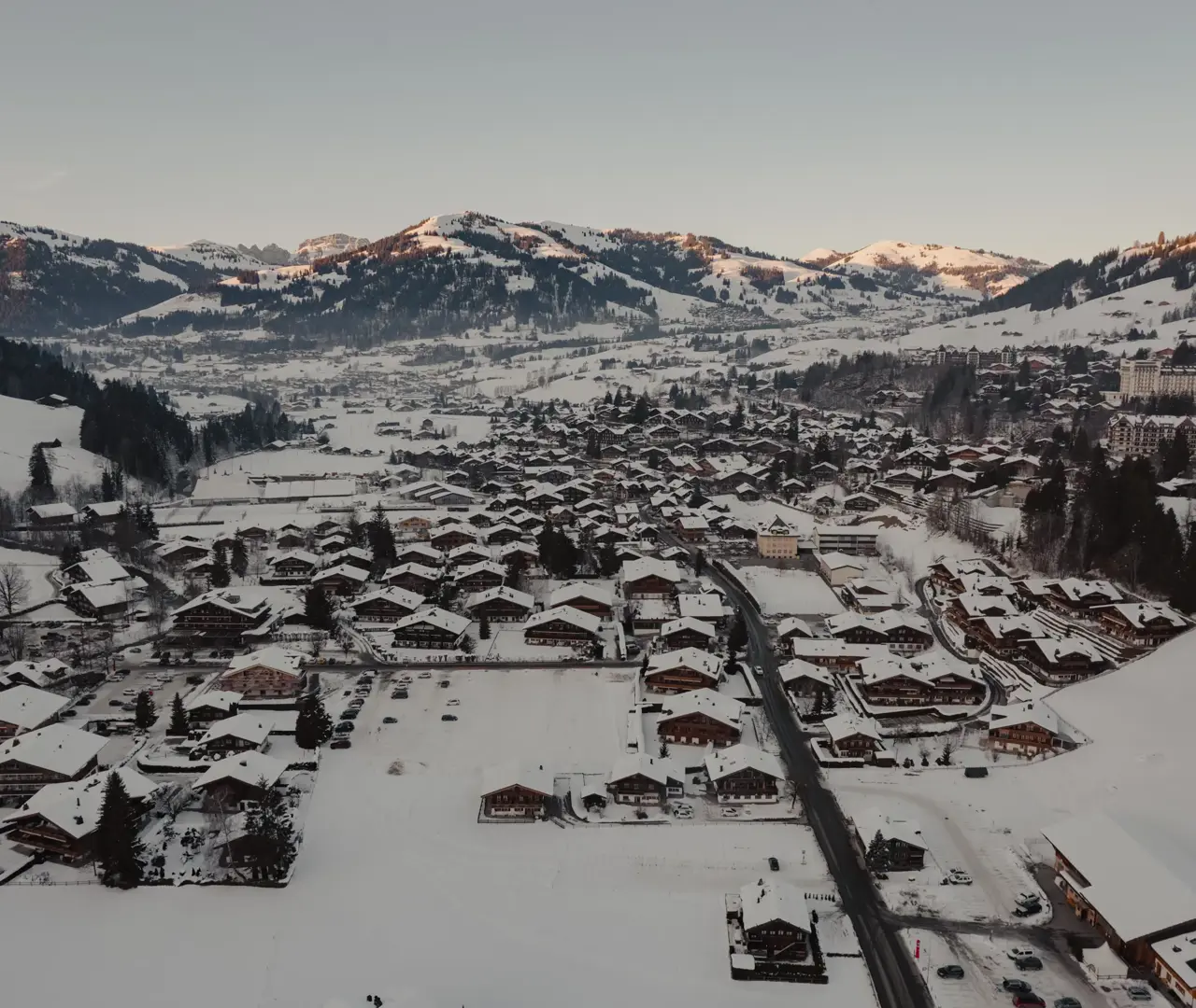 Вид с воздуха на заснеженное альпийское поселение, окружённое горами на рассвете, с деревянными шале и извилистыми дорогами в долине.