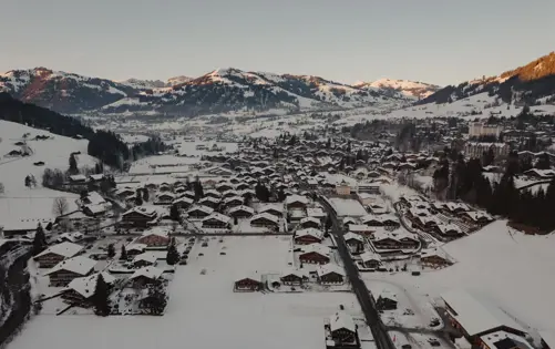 Vue aérienne d’un village alpin enneigé entouré de montagnes au lever du soleil, avec des chalets en bois et des routes sinueuses nichées dans la vallée.