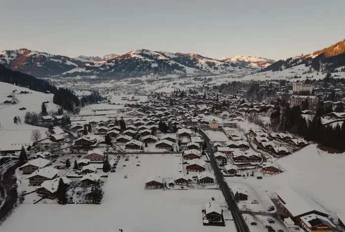 Vue aérienne d’un village alpin enneigé entouré de montagnes au lever du soleil, avec des chalets en bois et des routes sinueuses nichées dans la vallée.