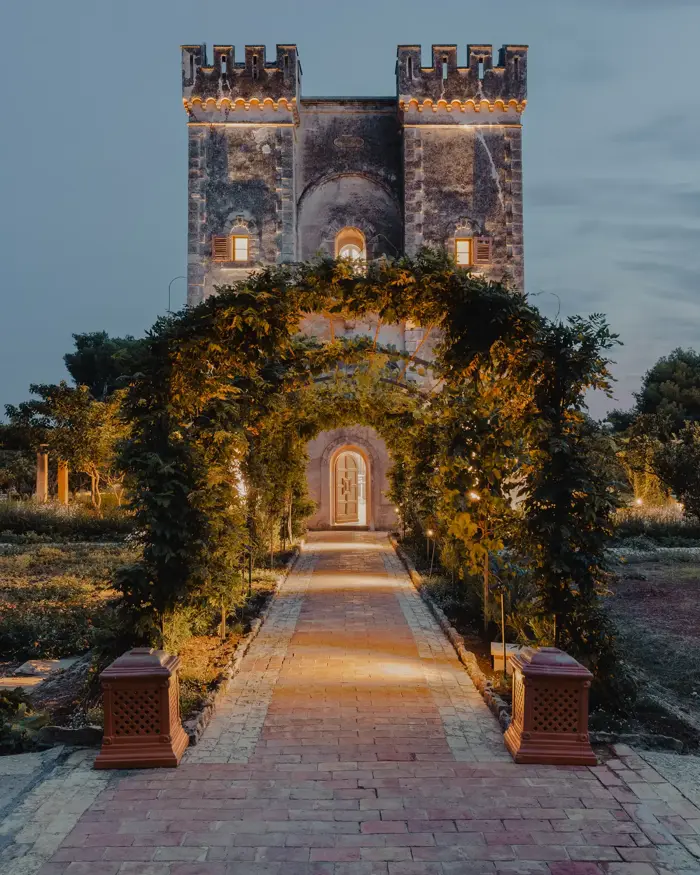Villa historique en pierre avec deux tours, encadrée par une arche de jardin illuminée au crépuscule, entourée de verdure et d’arbres fruitiers.