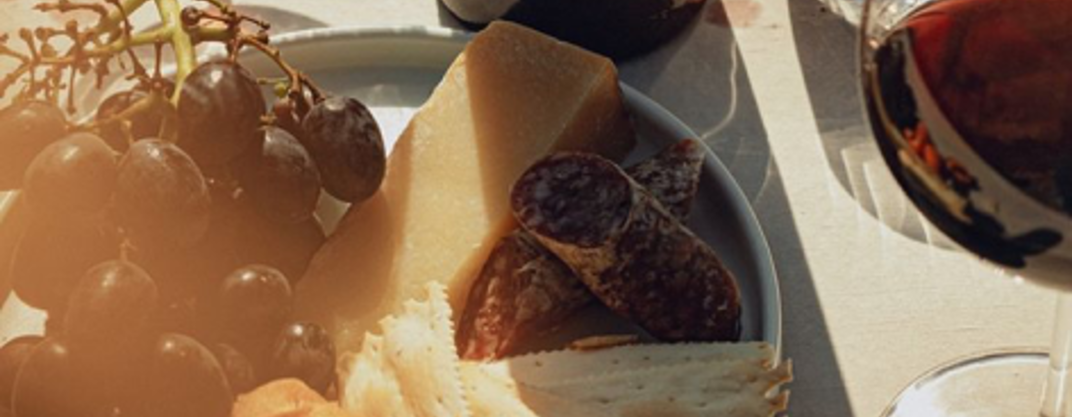 Charcuterie plate with assorted cheeses, grapes, and cured meats paired with a glass of red wine in warm sunlight.
